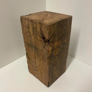 Puede incluir: Un bloque de madera rectangular con un acabado marrón natural, que presenta patrones de veta visibles, grietas verticales y un nudo prominente en la cara frontal. La superficie superior muestra anillos de crecimiento concéntricos.