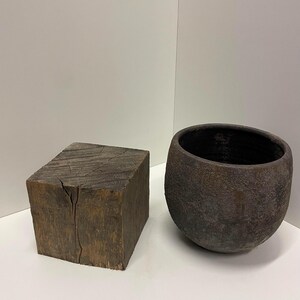 May include: A dark brown, weathered wooden cube with visible grain and a vertical crack on its front face, positioned next to a round, dark brown, textured ceramic bowl with a wide opening and a deep, dark interior cavity.