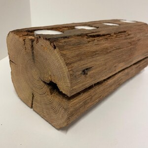 May include: A rustic wooden candle holder made from a section of a log. The brown log has four circular cutouts holding white tea light candles. The wood grain and natural imperfections are visible, adding to its charm.
