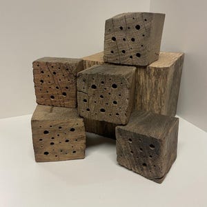 Peut inclure: Un ensemble de cinq blocs de bois rectangulaires et patinés, disposés en pile. Chaque bloc présente une texture naturelle et brute avec un grain de bois visible et de multiples trous circulaires percés sur les surfaces avant.