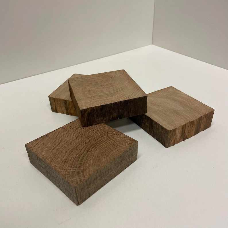 May include: Three square wooden coasters with visible grain patterns and natural brown tones. Each coaster features a thick, block-like construction with rough-hewn edges, showcasing the organic texture and rings of the wood material in a stack.