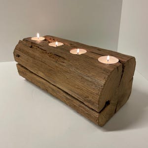 May include: A rustic wooden candle holder with four lit tea light candles. The candle holder is made from a section of a log, with four circular indentations to hold the candles. The wood has a natural, weathered appearance.
