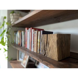 May include: A square block of weathered, light brown wood with a rough, textured grain sits on a dark wooden shelf. Behind the block, a row of various books with colorful spines is arranged vertically, creating a layered display on the shelf.