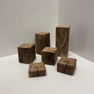 Op de afbeelding: Een verzameling van zes natuurlijke bruine houten blokken van verschillende hoogtes en breedtes. Elk blok heeft een zichtbare houtnerf en een duidelijke verticale scheur die door het midden van de bovenkant loopt, wat de ruwe textuur benadrukt.