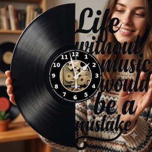 Op de afbeelding: Een wandklok gemaakt van een zwarte vinylplaat met de tekst Life without music would be a mistake. De wijzerplaat heeft een centraal ontwerp met zichtbare metalen tandwielen, witte cijfers en dunne witte wijzers voor de tijd.
