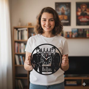 Puede incluir: Un reloj de pared circular en blanco y negro con una silueta de The Beatles. El reloj tiene números romanos y manecillas, con el nombre de la banda en la parte inferior. El reloj lo sostiene una persona que lleva una camiseta blanca.