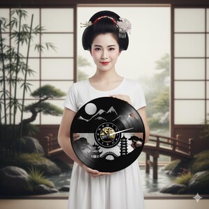 Könnte beinhalten: Eine schwarze Vinyl-Schallplattenuhr mit einem japanischen Landschaftsdesign mit Bergen, einer Brücke und einer Pagode. Die Uhr hat ein goldfarbenes Zifferblatt und weiße Zeiger. Die Uhr wird von einer Person in einem weißen Kleid gehalten.