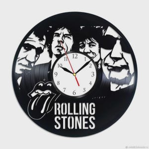 Puede incluir: Reloj de vinilo negro con las caras de los miembros de la banda Rolling Stones y el icónico logo de la lengua. La esfera del reloj es blanca con números y manecillas negras, y un segundero rojo. Las palabras "ROLLING STONES" están en blanco.