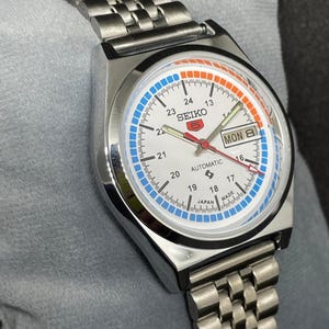 Puede incluir: Un reloj de pulsera autom&aacute;tico Seiko 5 plateado con esfera blanca. El reloj presenta detalles en azul y naranja, visualizaci&oacute;n de d&iacute;a y fecha, y una correa de acero inoxidable. La esfera muestra el texto "SEIKO 5 AUTOMATIC JAPAN MADE".