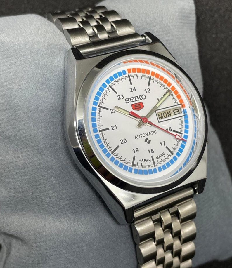 Puede incluir: Un reloj de pulsera autom&aacute;tico Seiko 5 plateado con esfera blanca. El reloj presenta detalles en azul y naranja, visualizaci&oacute;n de d&iacute;a y fecha, y una correa de acero inoxidable. La esfera muestra el texto "SEIKO 5 AUTOMATIC JAPAN MADE".
