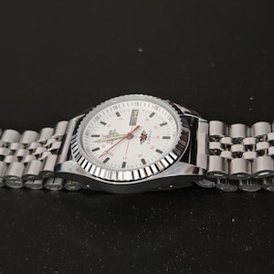 Puede incluir: Un reloj de pulsera plateado con esfera blanca y correa plateada. El reloj tiene una esfera redonda con marcadores de hora negros y agujas rojas. La correa está hecha de pequeños eslabones metálicos conectados.