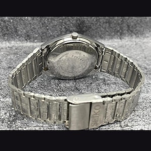Puede incluir: Un reloj de pulsera Seiko plateado con una correa de acero inoxidable. La esfera del reloj es visible, mostrando el nombre de la marca y otros detalles. La correa es de estilo eslabones con cierre de hebilla. El reloj es resistente al agua.
