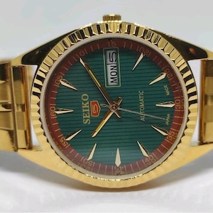 May include: A gold-toned Seiko 5 automatic wristwatch. The watch face is green with gold hands and markers, and a day-date display. The watch band is also gold-toned, with a textured design. The watch is marked "SEIKO 5 AUTOMATIC MADE IN JAPAN".