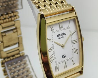 May include: A gold-toned Seiko Quartz watch with a rectangular face and a matching mesh band. The watch face is white with black Roman numerals and the Seiko Quartz logo. The watch is displayed on a clear stand.