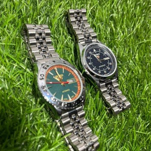 May include: Two stainless steel Seiko watches with metal straps are displayed on green grass. One watch features a green dial with orange detailing, the other a black dial. Both watches have a classic, sporty design.