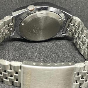 Puede incluir: Un reloj de pulsera plateado con una correa de metal. La esfera del reloj no es visible, pero la parte trasera de la caja del reloj es visible, con texto y marcas. La correa est&aacute; hecha de peque&ntilde;os eslabones met&aacute;licos conectados.