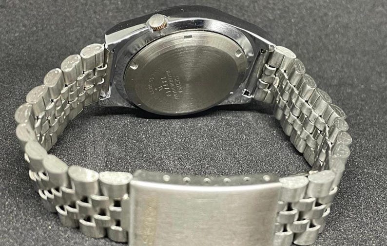 Puede incluir: Un reloj de pulsera plateado con una correa de metal. La esfera del reloj no es visible, pero la parte trasera de la caja del reloj es visible, con texto y marcas. La correa est&aacute; hecha de peque&ntilde;os eslabones met&aacute;licos conectados.