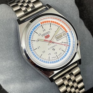 Puede incluir: Un reloj de pulsera autom&aacute;tico Seiko 5 plateado con esfera blanca. El reloj presenta una pantalla de d&iacute;a y fecha, detalles en azul y naranja, y una correa de acero inoxidable. La esfera muestra el texto "SEIKO 5 AUTOMATIC" y "MON 8".