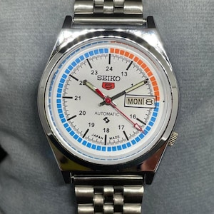 Puede incluir: Un reloj de pulsera autom&aacute;tico Seiko 5 plateado con esfera blanca. El reloj presenta detalles en azul y naranja, visualizaci&oacute;n de d&iacute;a y fecha, y una correa de acero inoxidable. La esfera muestra el texto "SEIKO 5 AUTOMATIC JAPAN MADE".