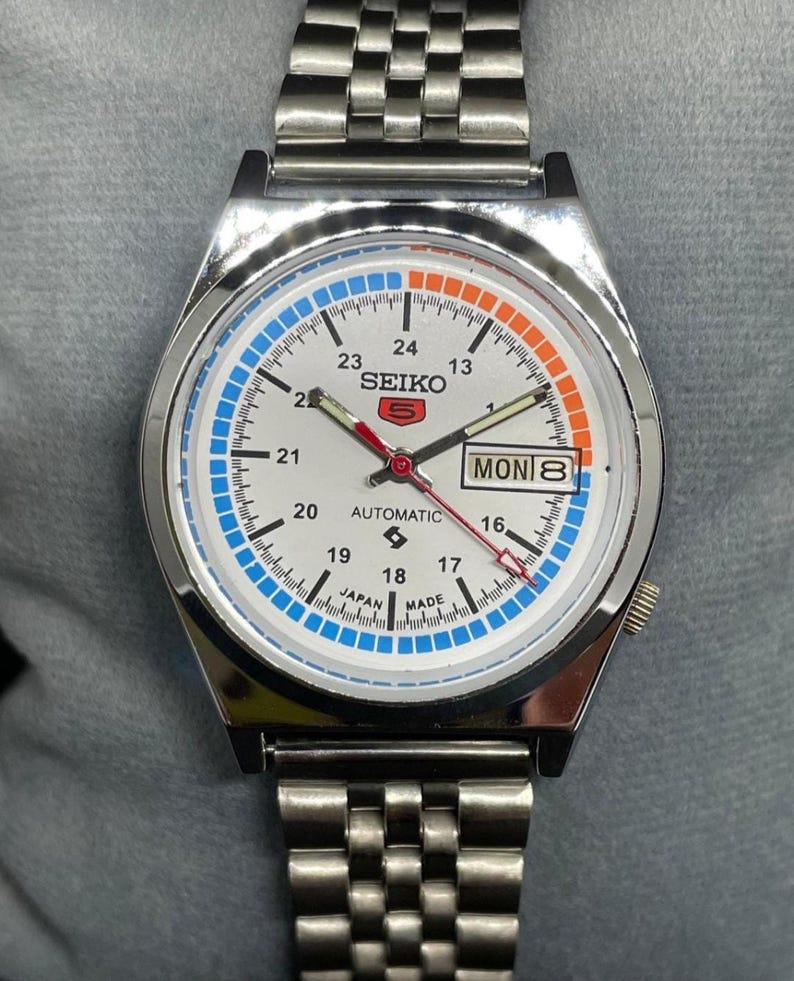 Puede incluir: Un reloj de pulsera autom&aacute;tico Seiko 5 plateado con esfera blanca. El reloj presenta detalles en azul y naranja, visualizaci&oacute;n de d&iacute;a y fecha, y una correa de acero inoxidable. La esfera muestra el texto "SEIKO 5 AUTOMATIC JAPAN MADE".
