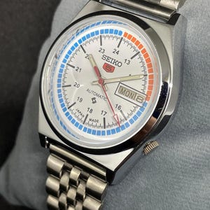 Puede incluir: Un reloj de pulsera autom&aacute;tico Seiko 5 plateado con esfera blanca, con detalles en azul y naranja. El reloj tiene una pantalla de d&iacute;a y fecha y una correa de acero inoxidable. La esfera del reloj muestra el texto "SEIKO 5 AUTOMATIC" y "MON 8".