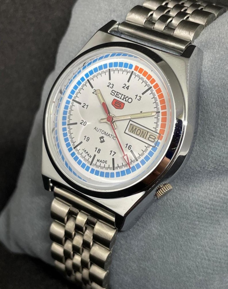 Puede incluir: Un reloj de pulsera autom&aacute;tico Seiko 5 plateado con esfera blanca, con detalles en azul y naranja. El reloj tiene una pantalla de d&iacute;a y fecha y una correa de acero inoxidable. La esfera del reloj muestra el texto "SEIKO 5 AUTOMATIC" y "MON 8".