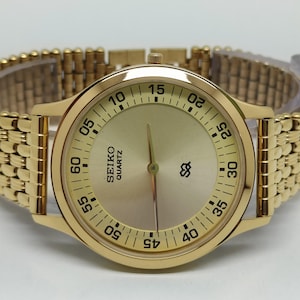 May include: Gold-toned Seiko Quartz watch with a gold band. The watch face is gold with black numerals and hands. The band is a flexible, woven style. The watch is displayed on a clear plastic stand.