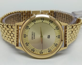 May include: Gold-toned Seiko Quartz watch with a gold band. The watch face is gold with black numerals and hands. The band is a flexible, woven style. The watch is displayed on a clear plastic stand.
