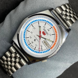 Puede incluir: Un reloj de pulsera autom&aacute;tico Seiko plateado con esfera blanca, con detalles en azul y naranja. El reloj tiene una pantalla de fecha y una correa de metal. La esfera muestra la palabra "SEIKO" y "AUTOMATIC".