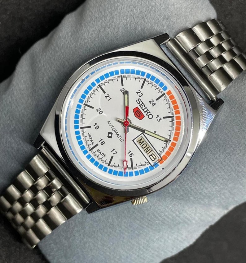 Puede incluir: Un reloj de pulsera autom&aacute;tico Seiko plateado con esfera blanca, con detalles en azul y naranja. El reloj tiene una pantalla de fecha y una correa de metal. La esfera muestra la palabra "SEIKO" y "AUTOMATIC".