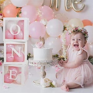 Pode incluir: Uma menina com um vestido rosa e uma coroa de flores sorri em frente a uma decoração de aniversário. A cena inclui um bolo branco em um suporte decorativo, balões e caixas que soletram "ONE" cheias de balões rosa.