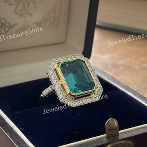 Könnte beinhalten: Ein Ring mit einem rechteckigen Smaragd-Edelstein, umgeben von Gold und Diamanten, präsentiert in einer offenen, dunklen Holzschatulle mit blauem Samt. Der Smaragd ist ein leuchtendes Grün.