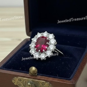 Peut inclure: Une bague en rubis et diamants dans un écrin en bois. La bague présente un grand rubis de taille ovale entouré d'un halo de petits diamants ronds. La boîte est marron foncé avec des ferrures en laiton et doublée de velours bleu foncé.