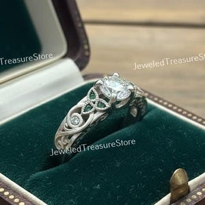May include: A silver ring with a round, clear center stone and smaller green and clear stones. The ring is in a dark brown jewelry box with a green velvet interior. The box is labeled "Jeweled Treasure Store".
