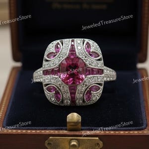 May include: A square-shaped, silver-toned ring with a large, round, pink gemstone at the center. The ring is surrounded by smaller pink stones and clear crystals. The ring is displayed in a wooden jewelry box with a blue interior.