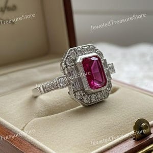 Puede incluir: Un anillo de plata con una piedra preciosa rectangular de color rosa intenso en el centro, rodeada de piedras más pequeñas y transparentes. El anillo tiene un diseño geométrico y se muestra en un joyero de madera.