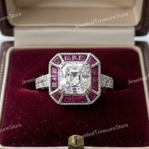 Könnte beinhalten: Ein silberner Ring mit einem großen, quadratisch geschliffenen Diamanten, umgeben von kleinen, roten Edelsteinen. Der Ring wird in einer offenen, bordeauxfarbenen Schmuckschatulle präsentiert. Das Design ist aufwendig.