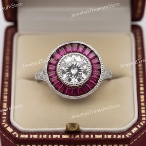 Puede incluir: Un anillo de estilo vintage con una piedra central grande, redonda y transparente, rodeada por un halo de piedras rectangulares de color rojo intenso. El anillo está engarzado en una banda plateada, presentado en un joyero burdeos.