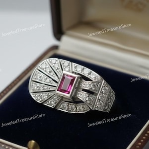 May include: A silver ring with a rectangular, pink gemstone at the center, surrounded by smaller, clear stones. The ring has an Art Deco design, set in a dark blue velvet-lined box. The ring is from Jeweled Treasure Store.
