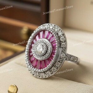 May include: An oval-shaped, silver-toned ring with a central design of vibrant pink baguette-cut stones radiating outwards. The ring is encircled by a row of small, clear stones, with additional detailing on the band. The ring is displayed in a jewelry box.