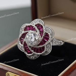 May include: A silver ring with a flower design. The ring has a central clear, round stone, encircled by smaller clear stones. The petals are made of rectangular, deep red stones, creating a striking contrast.
