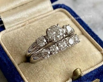 May include: A two-piece diamond ring set in a blue velvet ring box. The engagement ring features a large center stone flanked by smaller diamonds. The matching band has multiple smaller diamonds. The box is open, revealing the rings.