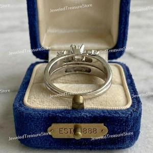 May include: A silver-colored ring set with a central, clear gemstone and smaller side stones, displayed in a blue velvet ring box. The box has a gold-colored plaque that reads "EST 888".