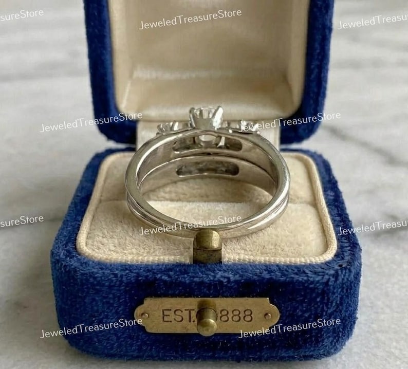 May include: A silver-colored ring set with a central, clear gemstone and smaller side stones, displayed in a blue velvet ring box. The box has a gold-colored plaque that reads "EST 888".