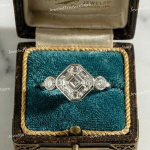 Puede incluir: Anillo de platino con un gran diamante central de talla cuadrada y dos diamantes redondos más pequeños a cada lado. El anillo se exhibe en un joyero vintage con interior de terciopelo verde azulado y ribete dorado.