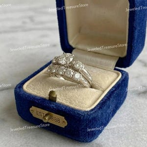 May include: A diamond engagement ring set in a blue velvet ring box. The set includes a ring with a large center stone and a band with smaller stones. The box has a gold clasp and the text "EST 888".