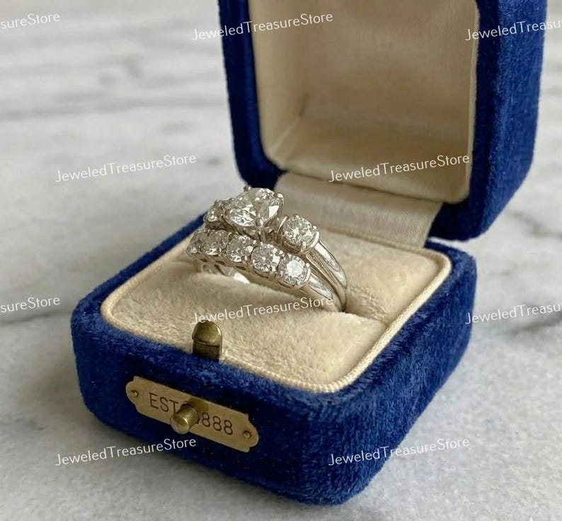 May include: A diamond engagement ring set in a blue velvet ring box. The set includes a ring with a large center stone and a band with smaller stones. The box has a gold clasp and the text "EST 888".