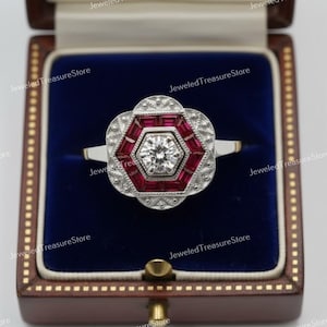 Peut inclure: Une bague de style vintage avec une pierre centrale claire et ronde, entourée d'un motif géométrique rouge et blanc. La bague est sertie dans un anneau en or et présentée dans un écrin en velours bleu.