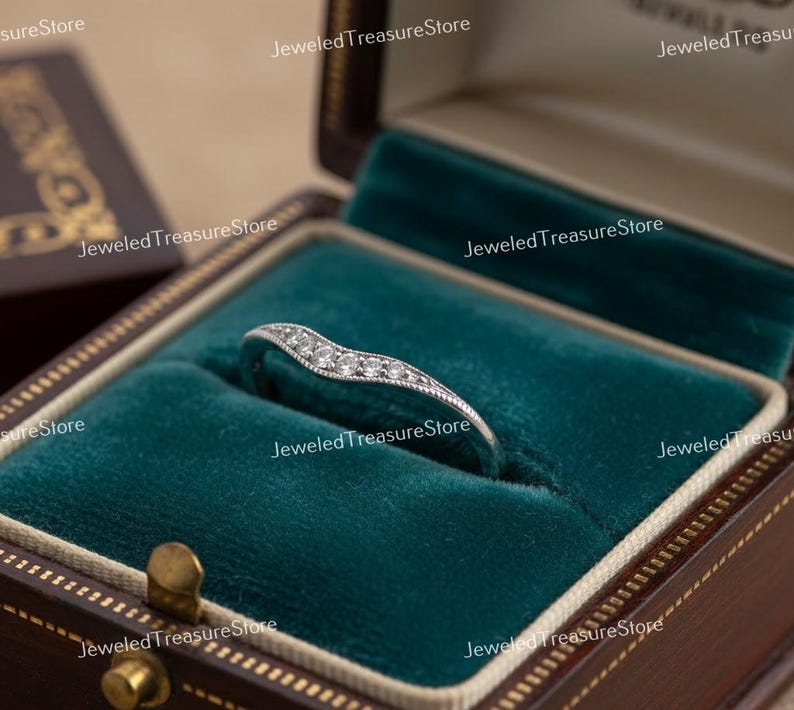 K&ouml;nnte beinhalten: Ein silberner Ring mit geschwungener Form, besetzt mit kleinen, klaren Edelsteinen. Der Ring wird in einer ge&ouml;ffneten, braunen Schmuckschatulle im Vintage-Stil mit tealfarbenem Samtfutter pr&auml;sentiert.