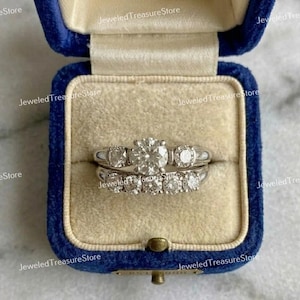 May include: A set of two diamond rings in a blue velvet ring box. The top ring has a large, round center stone flanked by two smaller stones. The bottom ring has a row of smaller diamonds. The box is open.